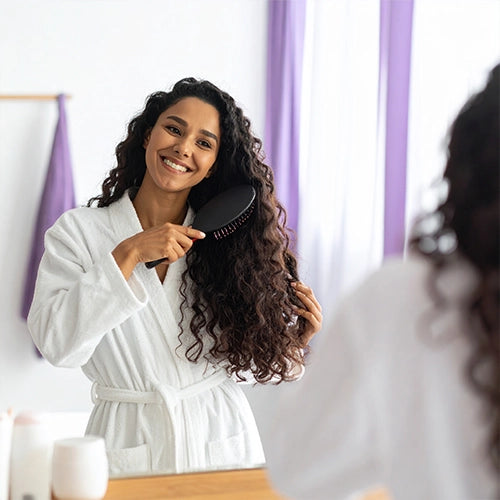 Woman in a white robe brushing her hair in front of a mirror with purple curtains.